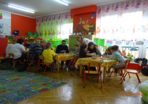 Dzieci i rodzice siedzą przy stolikach z ozdobami wielkanocnymi.